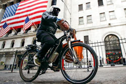 A delivery worker rides his electric bicycle past the New York Stock Exchange, March 16, 2020, in New York. New York City's thousands of food delivery workers will get places to recharge electric bike batteries, cellphones and themselves, under a city plan announced Monday, Oct. 3, 2022, to turn abandoned newsstands and other unused structures into facilities for the 'deliveristas.'