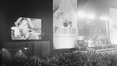 John Cougar Mellencamp performs both on stage and on one of two large screen television screens set up at the University of Illinois Memorial Stadium in Champaign, Ill. on Sept. 22, 1985 during the Farm Aid concert.