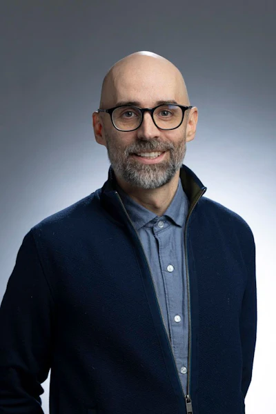 Professional headshot of bald man with beard wearing glasses and navy jacket over blue shirt