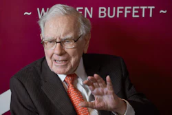 Elderly man in business suit and red tie speaking at event with burgundy background