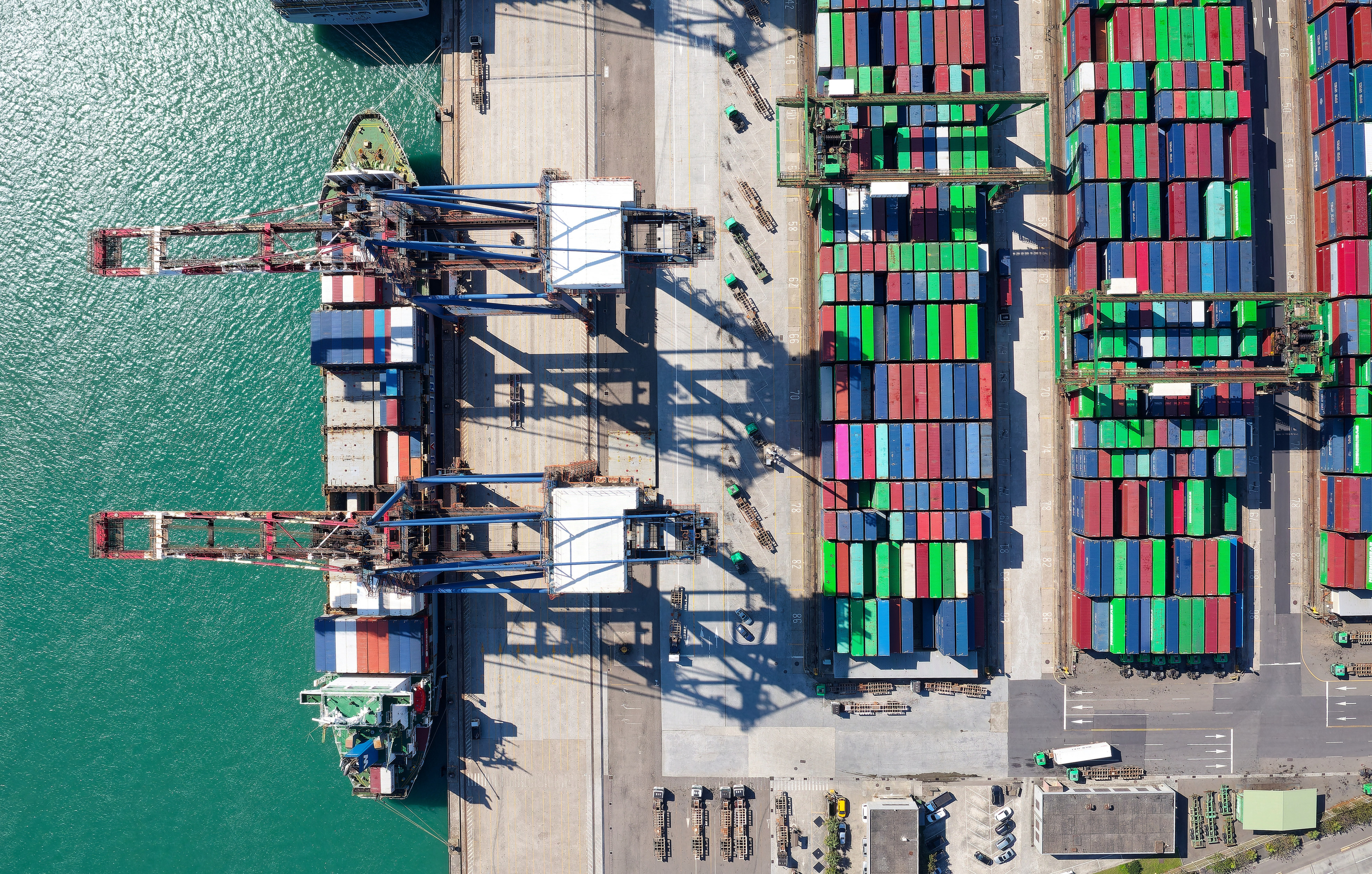 Aerial view of shipping port with cargo cranes, stacked containers, and docked vessel at waterfront terminal