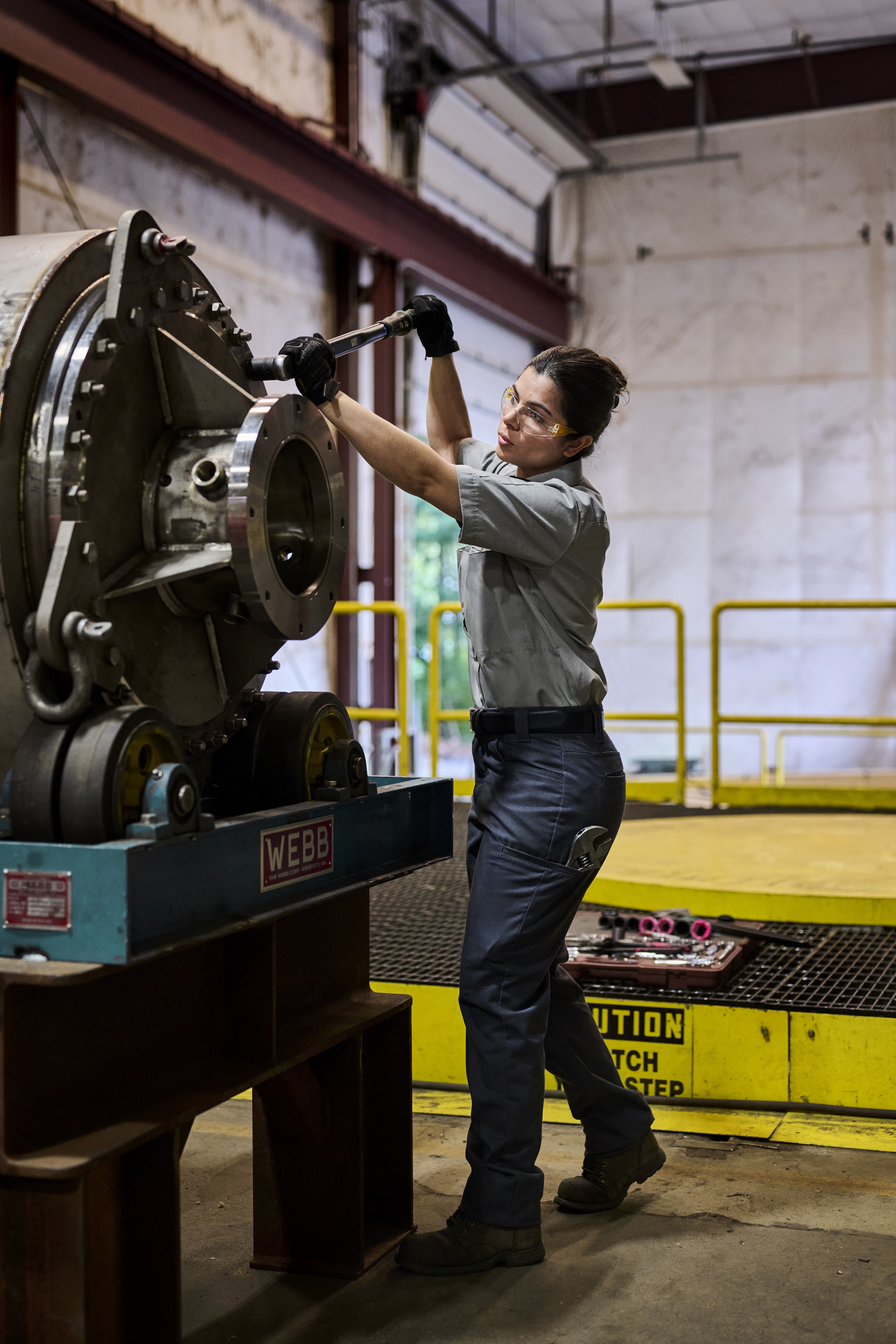 Female technician in safety glasses performing maintenance on industrial machinery in a factory warehouse.