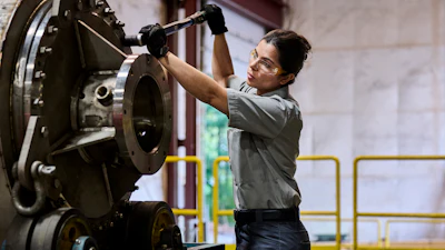 Female technician in safety glasses performing maintenance on industrial machinery in a factory warehouse.