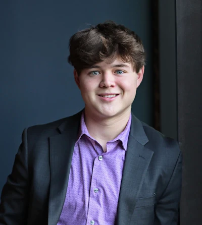 Professional headshot of young man wearing purple button-up shirt and dark blazer against blue background