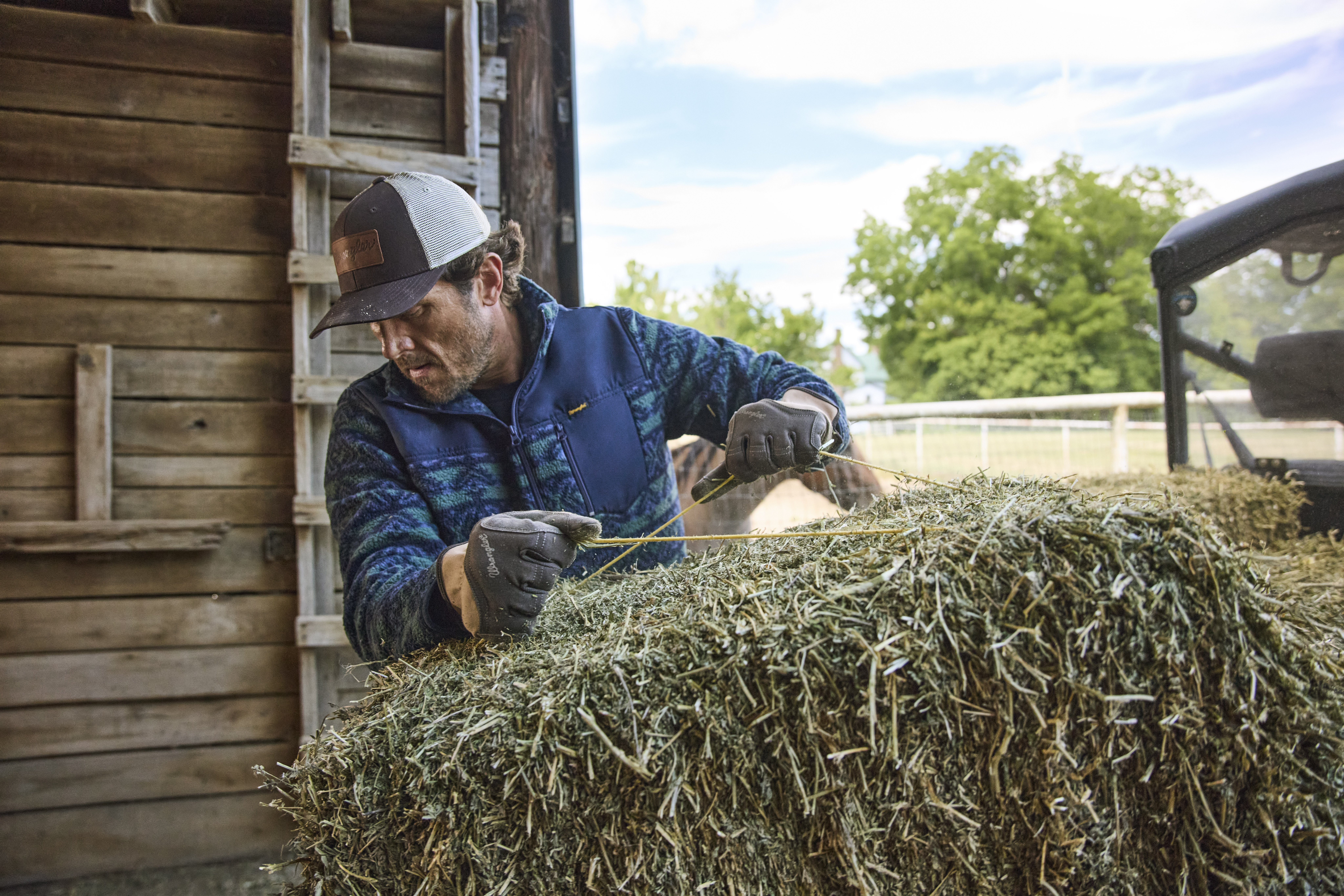 Farmer in blue jacket and cap handling hay bales near a wooden barn and truck