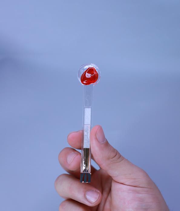 Hand holding a medical diagnostic test device with red collection sphere against blue background