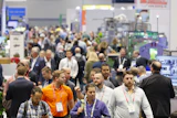 Crowded trade show floor with attendees wearing lanyards and ID badges networking among exhibition booths