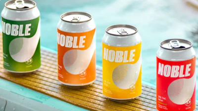 Four colorful Noble beverage cans in green, orange, yellow, and red arranged on a wooden surface near water