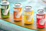 Four colorful Noble beverage cans in green, orange, yellow, and red arranged on a wooden surface near water