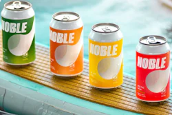 Four colorful Noble beverage cans in green, orange, yellow, and red arranged on a wooden surface near water