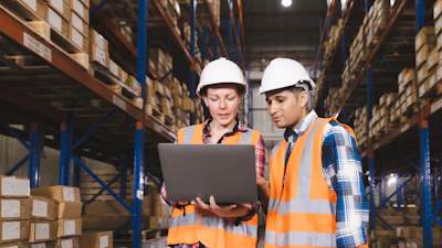 Two warehouse workers in safety gear reviewing inventory data on laptop in storage facility